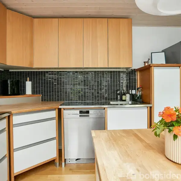 Et lyst køkken med træskabe og en mørk mosaikflisevæg. En opvaskemaskine er indbygget under køkkenbordet. En vase med orange blomster står på spisebordet.
