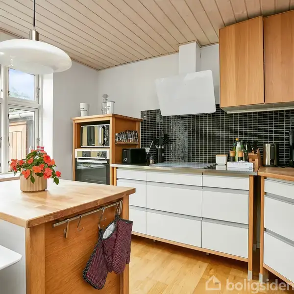 Køkken med hvide skabe og træborde, en ovn, mikrobølgeovn og diverse køkkenredskaber. Blomsterpotte på bordet, stor vinduessektion, hængende loftlampe. Lyse trægulve og sort flisebagvæg.