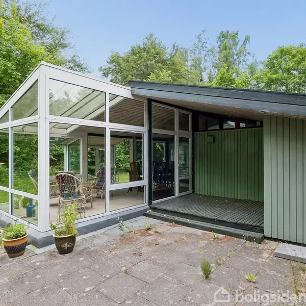 En moderne udestue med glasvægge står ved siden af et grønt træhus. Omkring er der en stenbelagt terrasse og frodig skovbevoksning, som skaber en fredfyldt atmosfære.