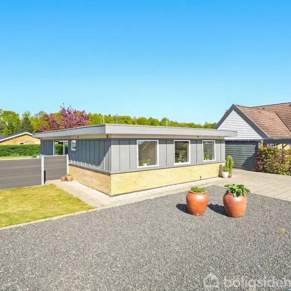 En flad tag bungalow står roligt på en grusvej, omgivet af en velholdt have og grønne buske, med en blå himmel over. To krukker står dekorativt foran huset.