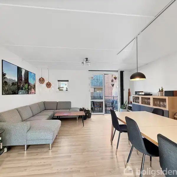 En stue med en grå hjørnesofa til venstre, et spisebord med stole til højre, og en dør til en balkon i baggrunden. Lyftelamper hænger over bordet.