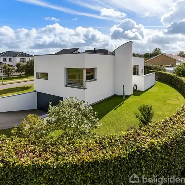 En moderne, hvid villa står omgivet af en veltrimmet hæk i et boligområde med enkelte træer og nabohuse under en skyet, blå himmel.