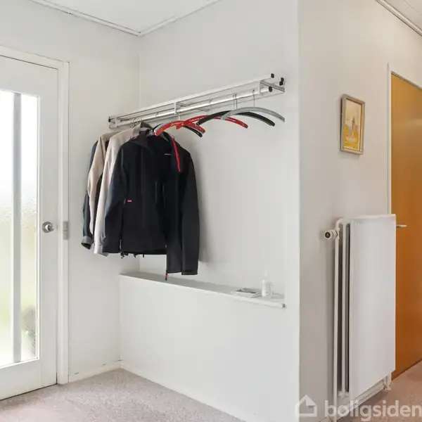 En række jakker hænger på en vægmonteret stang i en lys entre med tæppebelægning. Til venstre ses en glasdør med lodrette matterede striber, og til højre er en gul dør. En plante står i hjørnet.