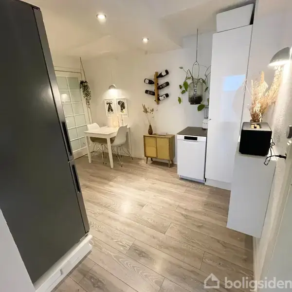 Et lille køkkenområde med moderne indretning. Et hvidt bord med to stole står i hjørnet under en lampe. Vægge har planter og vinholder. Lys trægulv skaber en varm atmosfære.