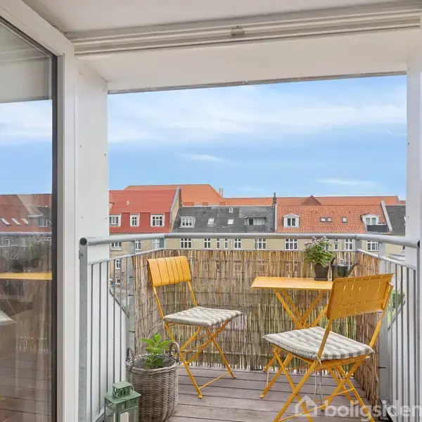 To gule stole og et bord står på en balkon, hvorfra man ser hustage og en blå himmel. Planterpotte til venstre.