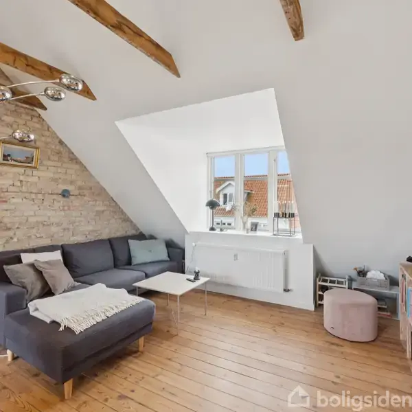 Sofaen står i et stuehjørne ved siden af en skråtstillet murstensvæg med en buet gulvlampe. Et fladt tv er modsat, mens et stort vindue giver udsigt til tagene.