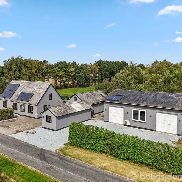 Et hus med solpaneler på taget står ved siden af en garagebygning med dobbeltporte. Omgivet af hække og træer, ligger ejendommen ved en stille vej i landlige omgivelser.