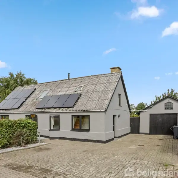 Et gråt hus med solpaneler på taget står stille i en gårdsplads med brosten. Omkring er der grønne buske og en garage. Himlen er blå med få skyer.