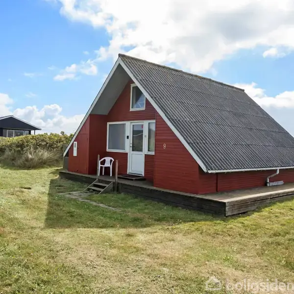 Rødt A-formet hus står på en græsplæne i solrigt vejr. Huset har en mørk tagflade og en træterrasse. Stol foran døren. Nummeret "9" er synligt.