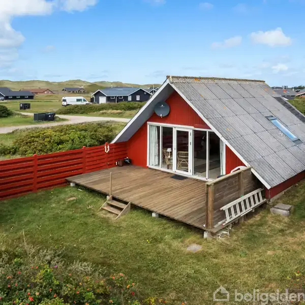 Et rødt, trekantet sommerhus med stråtag står på en græsplæne. Der er en træterrasse foran, omgivet af buske og flere huse i baggrunden under en blå himmel.