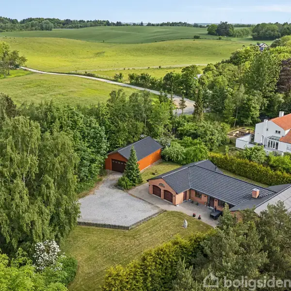 En moderne, brun murstensvilla med mørkt tag ligger omgivet af tæt grønt og åbne marker. Et separat rødt garageanlæg ses tæt ved, med en stor græsplæne og indkørsel foran.