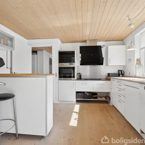 Et lyst køkken med hvide skabe og træloft indeholder en høj skammel ved køkkenøen. Sollys strømmer ind gennem store vinduer, der oplyser rummet og udsigten udenfor.