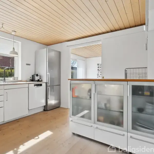 Et moderne køkken med hvide skabe og skuffer. Et rustfrit stål køleskab står i hjørnet. Store vinduer giver udsigt til en have. Flot træloft og trægulv.