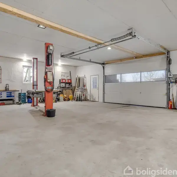 En stor garage med en bil-lift i midten, omgivet af værktøj og udstyr. Arbejdsborde langs væggene og en stor port i baggrunden.