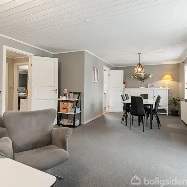Stue med gråt tæppegulv, sofa og lænestol til venstre, spisebord med sorte stole i midten, planter og lamper i det lyse rum.