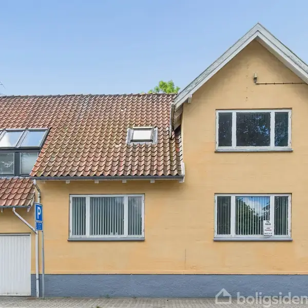 Gult hus med rødt tegltag har vinduer og en integreret garage. Der hænger et "Til salg"-skilt i vinduet. Det står på en gade med en parkeringsplads og tilstødende huse.