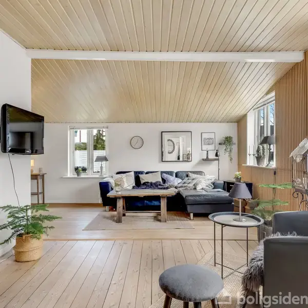 En lys stue har en mørk sofa arrangeret i hjørnet, pyntet med puder og tæpper. Væggene er dekoreret med billeder og en ur. Trægulve og træloft kompletterer den hyggelige atmosfære.