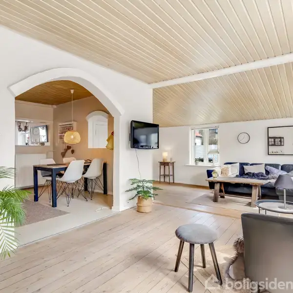 Et moderne, lyst opholdsrum med trægulv og loft, indeholder en sort sofa og et lille bord. I baggrunden ses et spiseområde med bord og stole under en pendel.