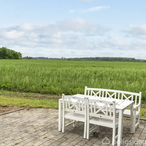 Hvide havemøbler står på en flisebelagt terrasse med udsigt over en stor grøn mark og træer i baggrunden under en let overskyet himmel.