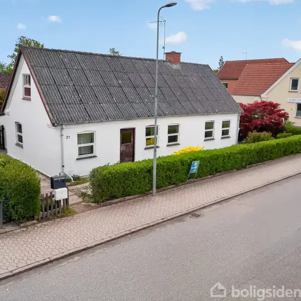 Et hvidt hus med sort tag og dør er omgivet af grøn hæk og beliggende langs en gade med fortov. Husnummeret "23" ses ved døren.