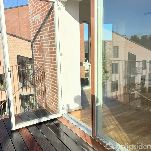 En glasdør åbner ud til en solrig altan med træplanker, omgivet af murede bygninger. Gitterrækværket kaster skygger på gulvet, og nabobygningerne kan ses i baggrunden.