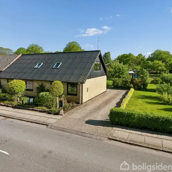 Hus med sort tag og beige facade står stille ved en sti omgivet af buske og træer. I baggrunden ses græsplæne, hække og flere grønne træer under en blå himmel.
