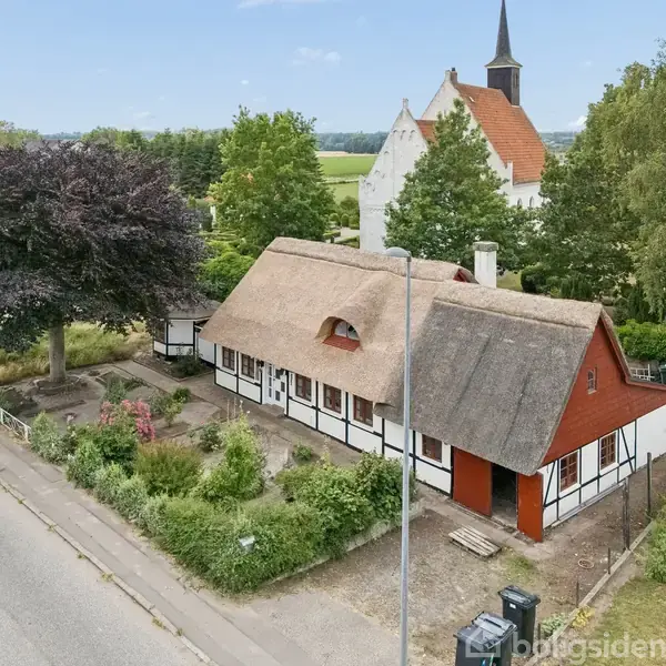 Bindingsværkshus med stråtag står stille ved en vej. Omgivet af have, stor træ og en kirke i baggrunden, ses huset i en landlig landskabsramme.