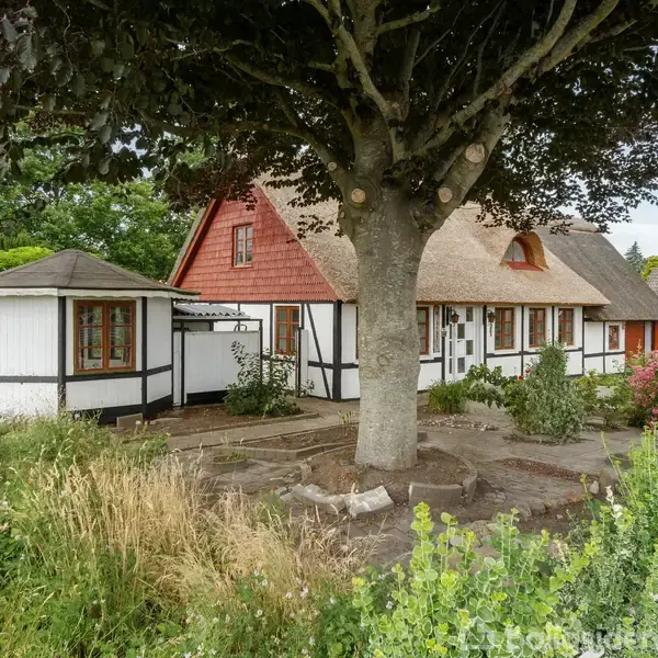 Et bindingsværkshus med stråtag og rød facade står i en frodig have med blomster og et stort træ i forgrunden. En pavillon ses til venstre.