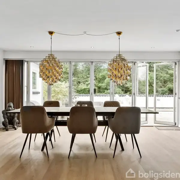 Et spisebord med seks stole står i en lys spisestue. To gyldne lamper hænger over bordet, og glasdøre åbner ud til en frodig have.