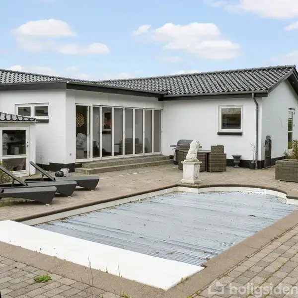 En hvid, moderne villa med sorte tagsten omkranser en terrasse med fliser. En overdækket swimmingpool ligger centralt, omgivet af liggestole og skulpturer. Skyfri, blå himmel ses i baggrunden.