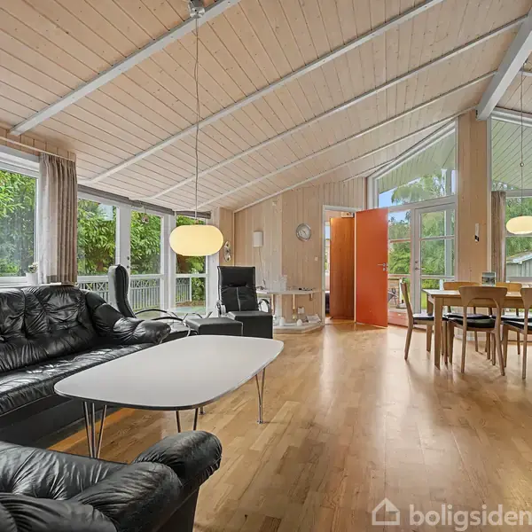 Sort lædersofa og to borde står i stue med trægulv og store vinduer. Spisebord med stole ses til højre. Grøn natur udenfor.