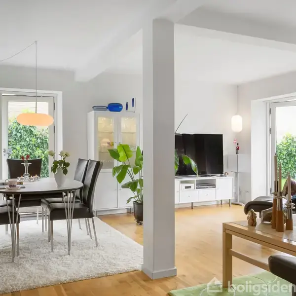 Spisestuemøbler står på et lys tæppe, mens en sort lædersofa og et TV optager stuen. Store vinduer og glasdøre åbner mod en grøn have.