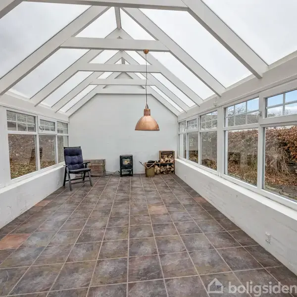 En lys vinterhave med flisegulv og skråt glasloft. Et par stole og brændekurve står i den ene ende. Store vinduer åbner udsigten til udendørs grønne områder.