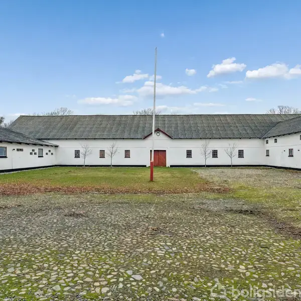 En hvid gårdbygning omkranser en gårdsplads med græs og grus. Midt i pladsen står en flagstang. Bygningen har små vinduer og lavt, gråt tag. Himlen er blå med skyer.
