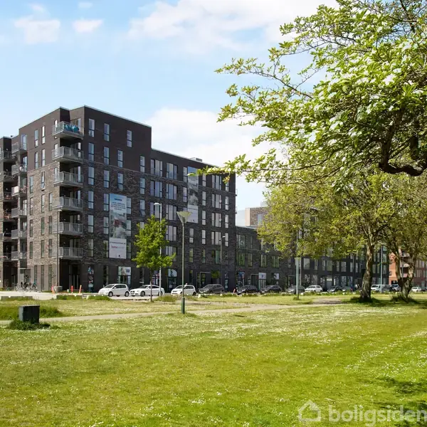 En moderne, mørk boligblok står stille i et bymiljø, omgivet af grønne træer og en åben græsplæne. I baggrunden ses flere parkerede biler langs gaden.
