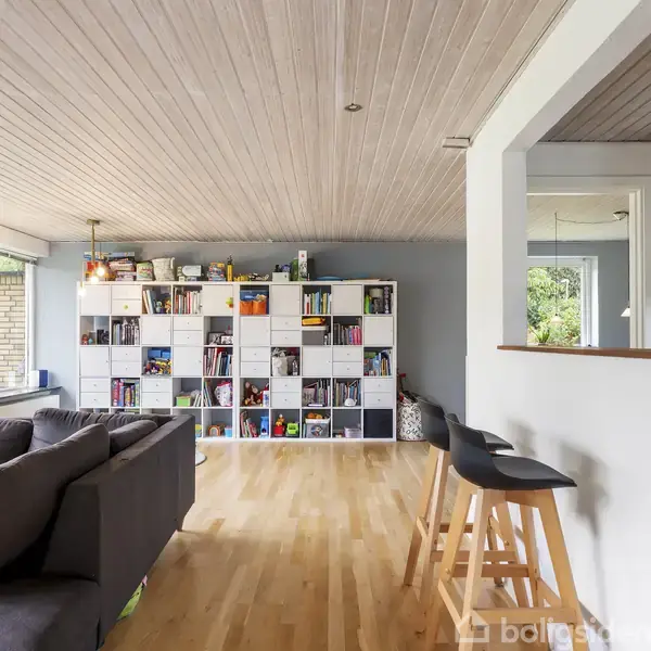 En stue med en grå sofa står ved siden af en stor reol fyldt med bøger og dekorative genstande. To barstole står ved en åbning mod køkkenet. Lys træparket og loft.