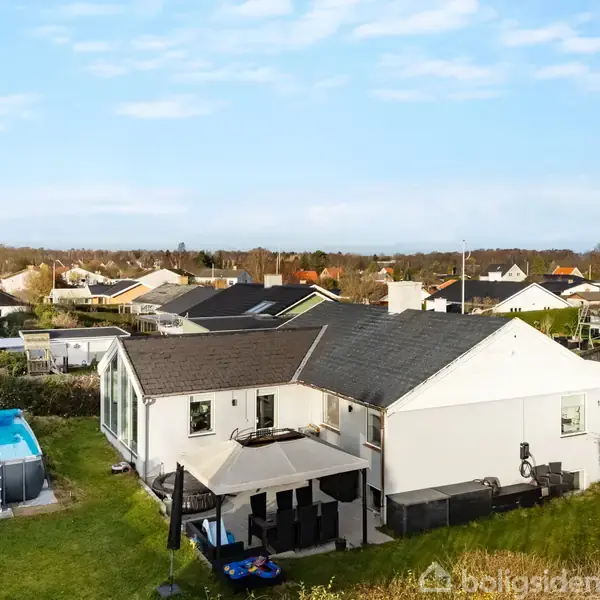 Et hvidt hus med gråt tag står i en have med en opstillede swimmingpool. Omkring huset er der andre hjem og grønt buskads under en blå himmel.