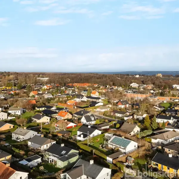 Boligområde med mange parcelhuse, der står stille i et naturskønt landskab. Husene har farverige tage, omgivet af velplejede haver og træer, under en klar blå himmel.