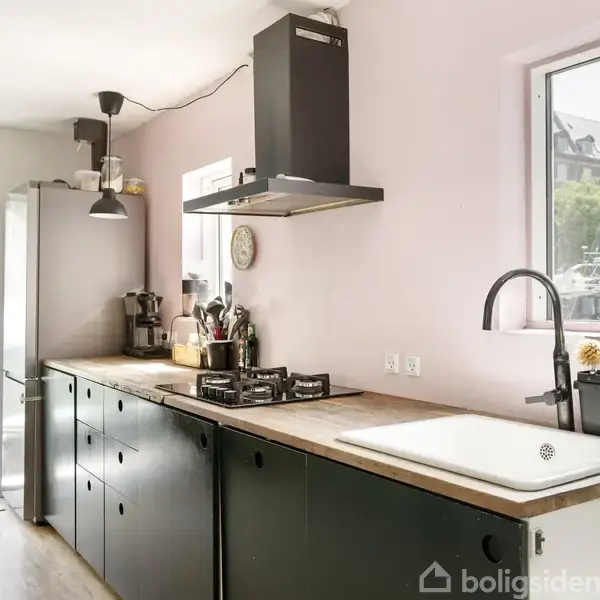 Sort køkken med gasblus og vask. Vinduet viser udsigt til både og bygninger. Dør fører ud til terrasse. Væggene er lyserøde, og der hænger en lampe over bordet.