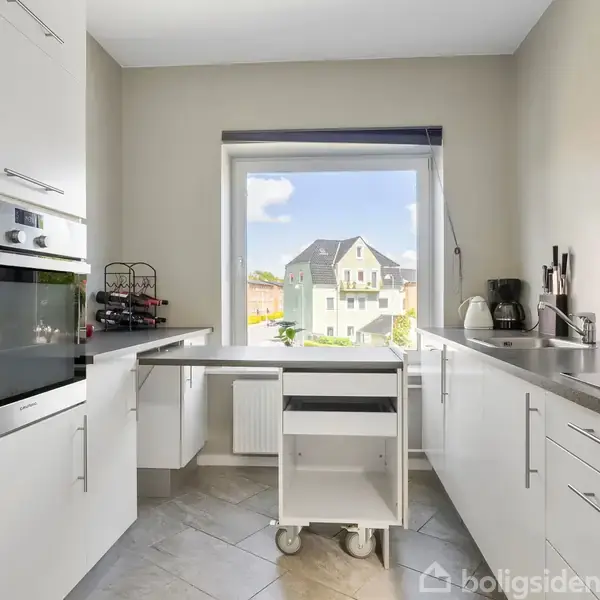 Et moderne køkken med hvide skabe og en indbygget ovn. En flytbar vogn står foran et stort vindue med udsigt til et hus og blå himmel udenfor.
