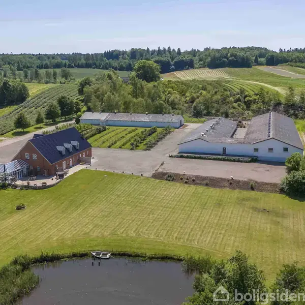En rød murstensejendom med drivhus står stille ved siden af landbrugsbygninger i et frodigt, kuperet landskab. En dam med en lille båd ses i forgrunden.