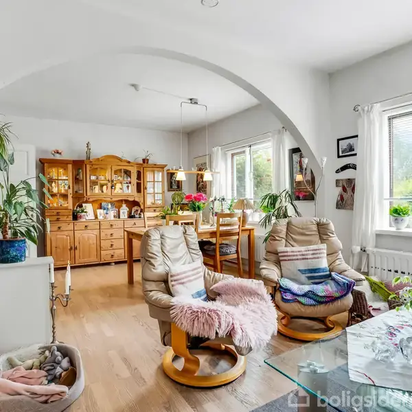 To lænestole med puder står foran et glasbord i en stue. Træskab fyldt med porcelæn ses i baggrunden, omgivet af planter ved vinduet, hvilket skaber en hyggelig atmosfære.