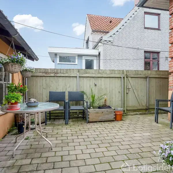 Terrasse med fliser. Et rundt bord og stole står ved siden af blomsterpotter og hængende planter. Trægærde og murstensvægge omkranser området, med huse i baggrunden.
