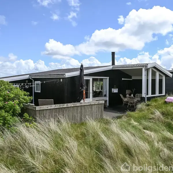 Et sort sommerhus med terrasse står blandt klitter og græs, under en blå himmel med spredte skyer. Terrassemøbler og en parasol ses på terrassen.