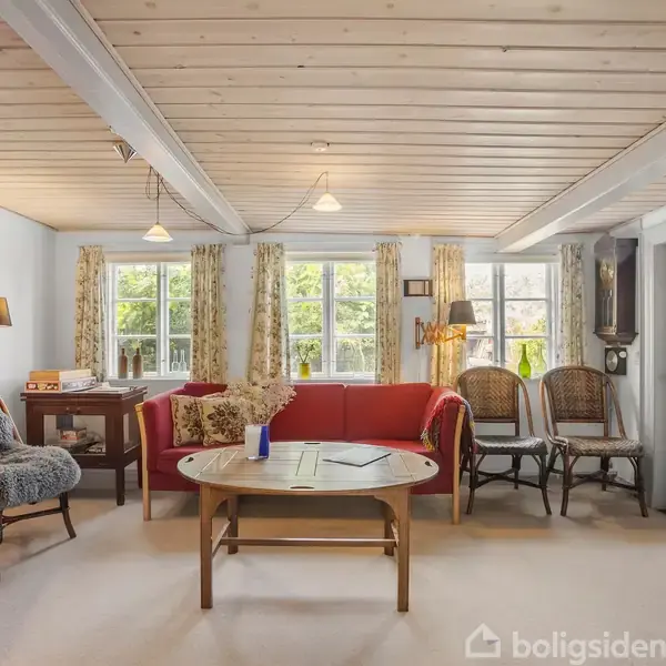 Stue med rød sofa foran tre vinduer, omgivet af kurvestole og lamper; et træbord står i midten, og der hænger et billede på væggen. Hvidt loft med synlige bjælker.