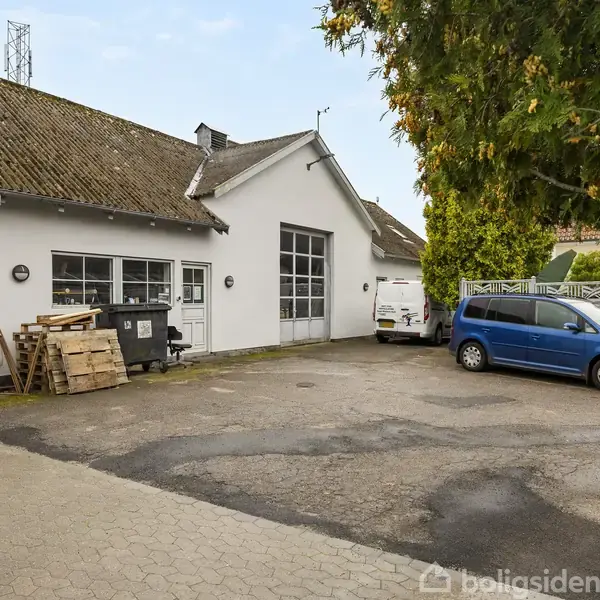 En hvid bygning med stor garageport, omgivet af en parkeringsplads. To biler, en hvid varevogn og en blå personbil, holder parkeret. På siden ses træpaller og containere.
