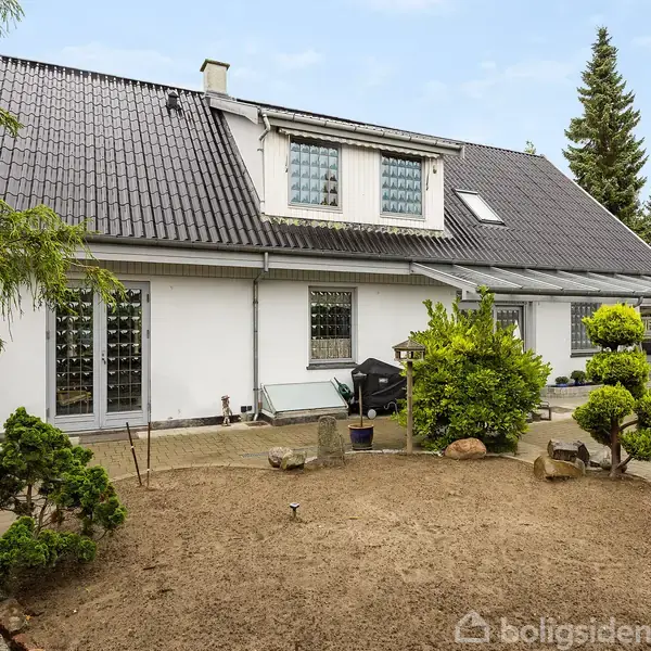 Et hvidt hus med skråt tag og store vinduespartier. Foran er en have med små træer, buske og sten. Omkringliggende fliser skaber en terrasse.