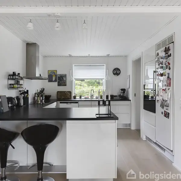 Et moderne køkken med en sort køkkenø, to sorte barstole og hvide vægge. Et vindue giver dagslys, og kunstværker hænger på væggene. Køleskabet er dækket med magneter og billeder.