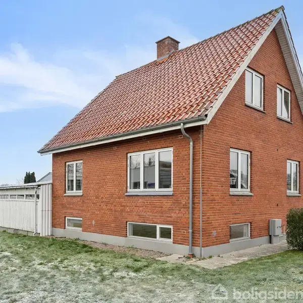 Rødt murstenshus med skråt tegltag ligger på en græsplæne. Huset har flere vinduer og en hvid garage på siden. Himlen er blå med lette skyer.