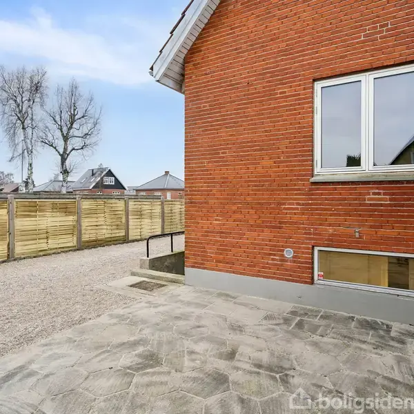 Rødt murstenshus med to vinduer, beliggende på flisebelagt gård. Et hegn af træ adskiller gården fra en grusvej. I baggrunden ses træer og flere tagrygge.